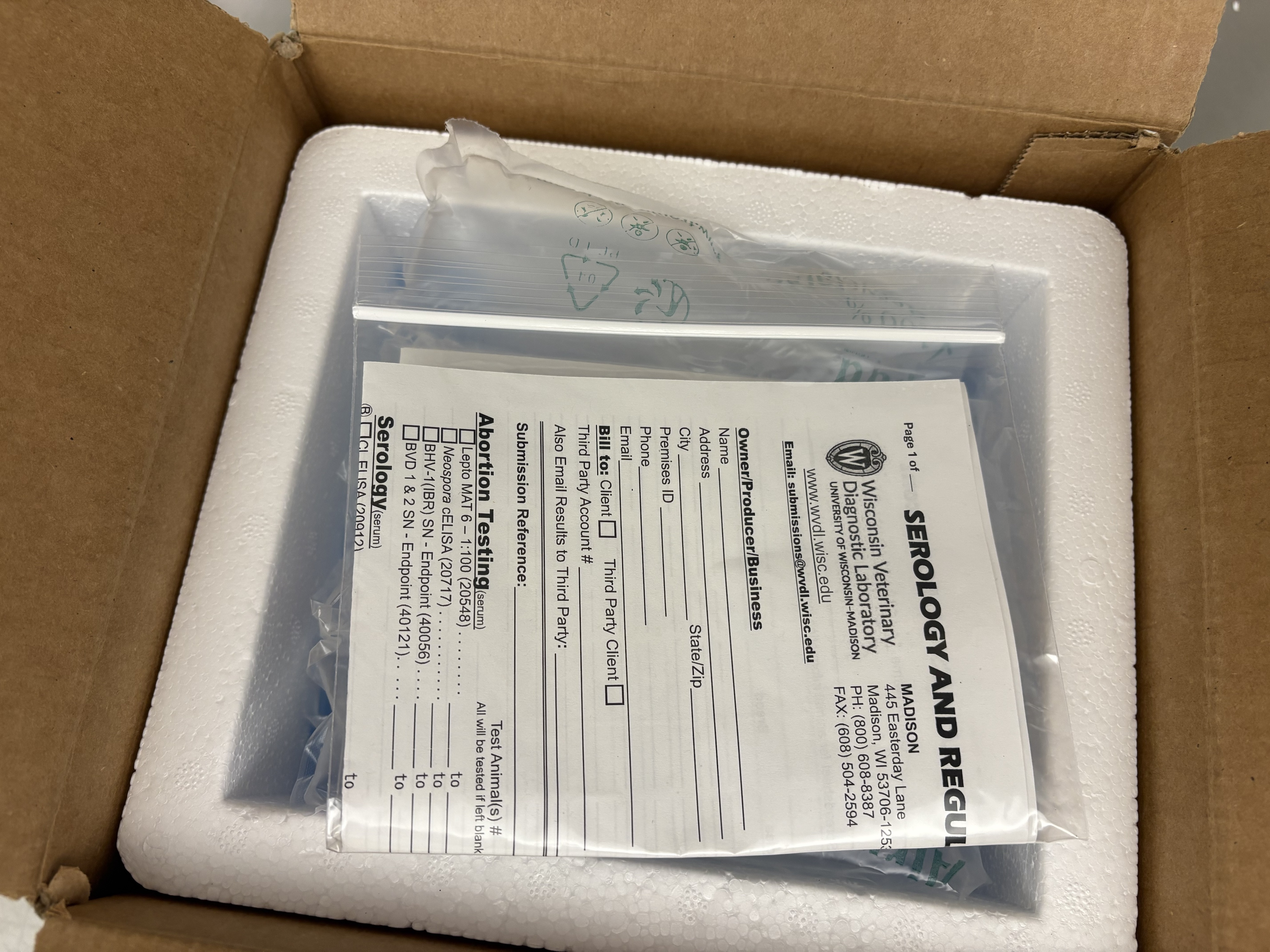 Sealed plastic bag containing submission paperwork placed flat on top of the inner foam lid inside the outer box.